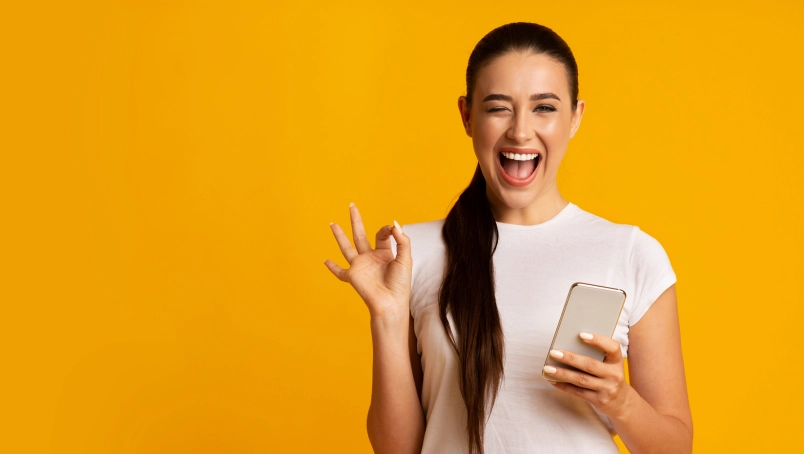 Glückliche Frau mit Smartphone vor gelbem Hintergrund, zeigt OK-Zeichen. Perfekt für digitale Inhalte.