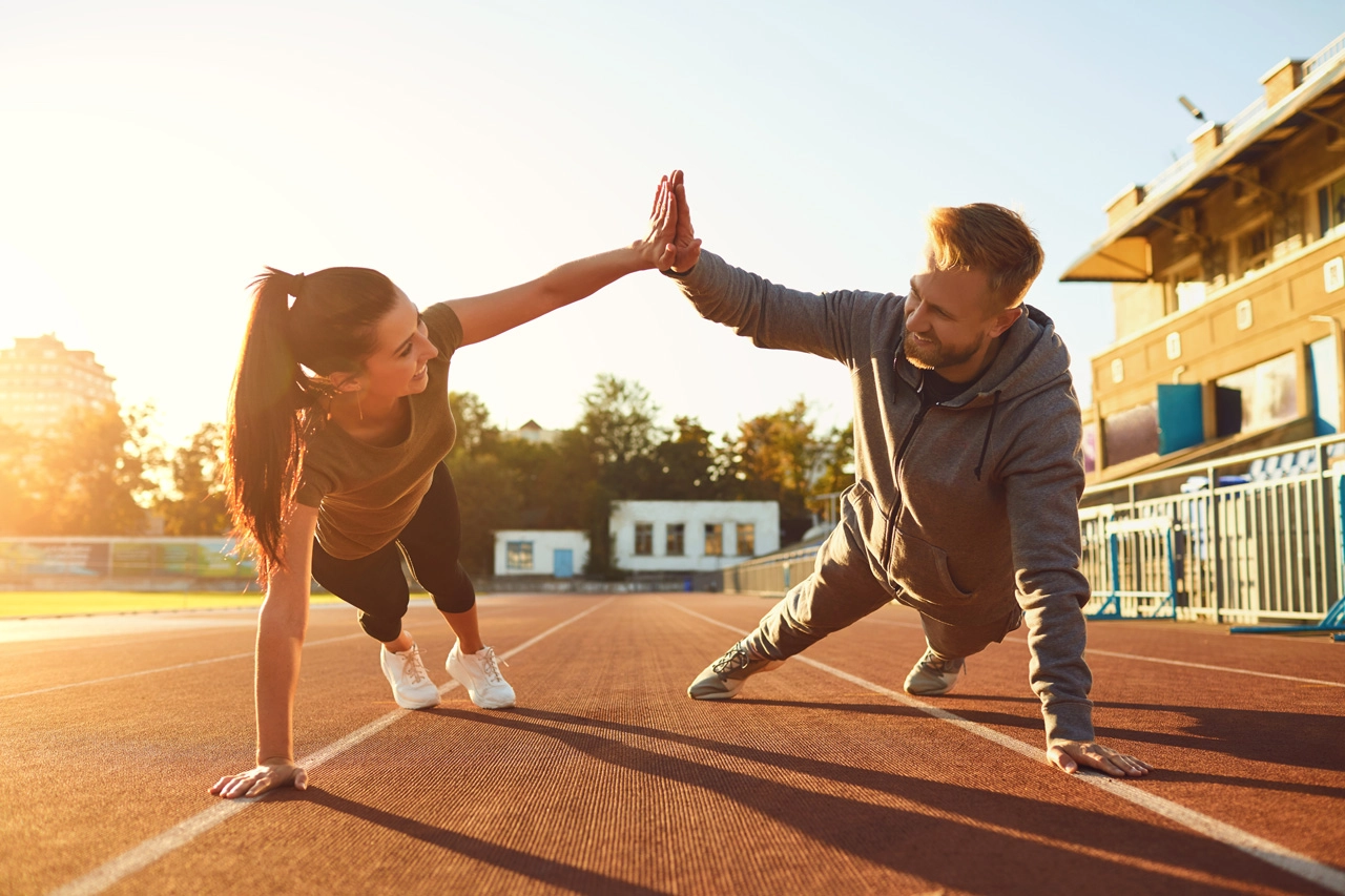 Fitness-Duo beim Plank-High-Five auf der Laufbahn bei Sonnenuntergang.